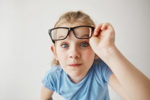 Close up portrait of cheerful small girl with blonde hair and blue eyes funny imitates adult person with glasses with surprised expression.