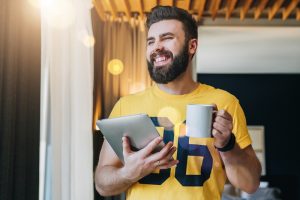 Young bearded man stands in room and holding tablet computer while drinking coffee. Guy freelancer working at home.