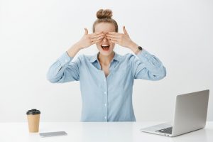 Portrait of amazed good-looking female entrepreneur in office, covering eyes with palms and waiting impatiently for surprise while sitting near laptop and smartphone, working over gray background