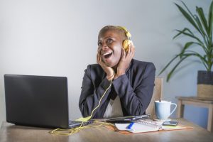 young happy black afro american woman listening to music with headphones excited and joyful working at laptop computer desk at modern workplace