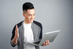 Excited happy asian man looking at laptop computer screen and celebrating the win isolated over gray background