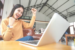 Young attractive asian woman looking at laptop computer feeling happy cheerful or excited expression success or win sitting on desk table in cafe coffee shop in wide close up view with soft tone.
