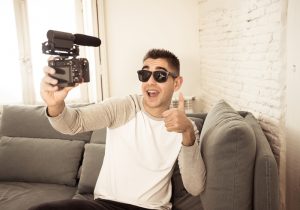 Young happy blogger recording video on camera for internet blog showing thumb up