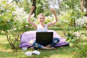 Happy freelancer working in the garden. Writing, surfing in the internet. Young woman relaxing and having fun in park area. Distance education, freelance concept.