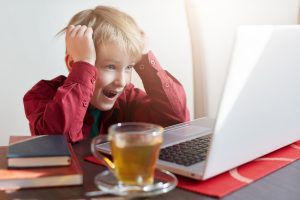 A surprised liitle boy with light hair dressed in red shirt sitting at the table and watching cartoons or video blogs online on his laptop and drinking hot tea enjoying leisure time